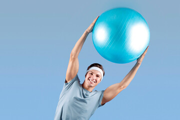 Positive young sportsman lifting fitness ball above his head, bending aside on lilac studio...