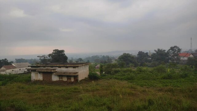 An abandoned building stands quietly on a grassy hill, surrounded by overgrown vegetation and distant houses, beneath a gloomy, overcast sky before dusk.