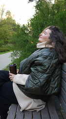 A young woman is sitting on a bench in a park with her eyes closed, dreaming. Holding a thermocup, she enjoys relaxing in the fresh air, recuperating and immersing herself in relaxation.