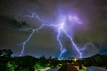 A powerful lightning bolt strikes over a suburban neighborhood at night, illuminating the rooftops...