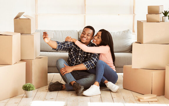 Positive black man and his wife moving to their own apartment together, taking selfie among boxes with belongings, panorama. African American couple making photo with smartphone on relocation day