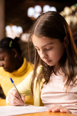 Diverse schoolgirls writing essays in a library in order to finish their homework assignments,...
