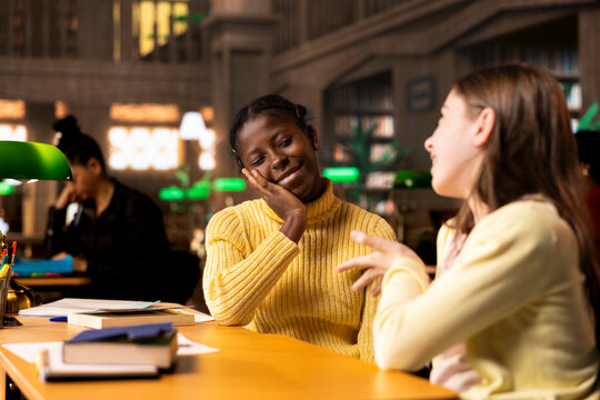 Happy two girls emphasizing their friendship with an embrace at the library, taking a break from study session and chatting about something. Classmates and friends having fun together.