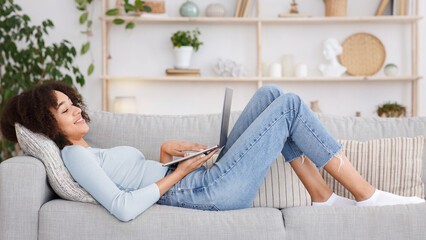 Fototapeta premium Relax with gadget, watching videos and chatting in social networks. Smiling young african american woman typing in laptop, writes message, lies on couch in living room interior, side view, free space