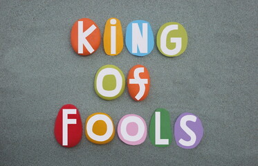 King of fools, creative funny logo composed with multi colored hand painted stone letters over green sand