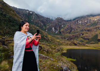 Naklejka premium Mujer quechua en vestimenta tradicional en América Latina, feliz mientras usa un teléfono inteligente mirada a un espacio publicidad y aislada en un fondo naturaleza,contactar,celular.Quechua