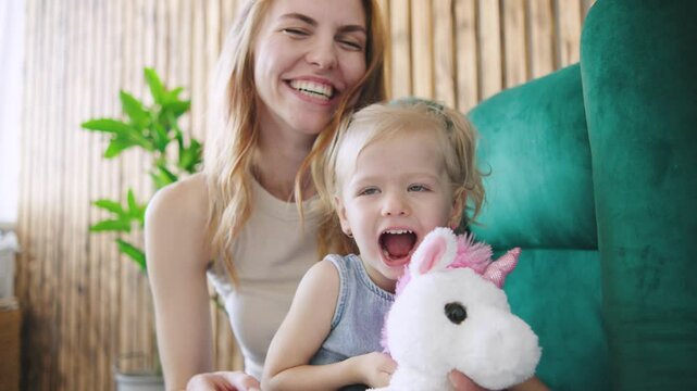 Cheerful mother and daughter playing together in a cozy indoor setting during the day, sharing joyful moments with a stuffed animal