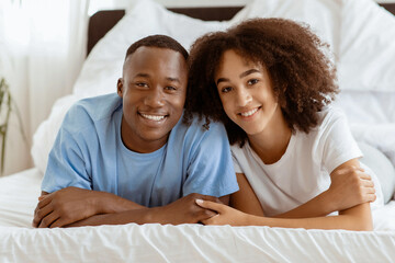 Young cute smiling couple together in bed, popular home bloggers during covid-19 lockdown. Happy millennial african american husband and wife looking at camera in cozy bedroom in morning, copy space