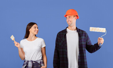 Happy Young Couple With Painting Tools In Hands Posing Over Blue Background. Millennial Spouses Holding Brushes, Ready For House Renovation And Improvement, Looking At Each Other And Smiling