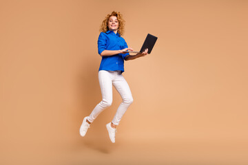 Confident young woman in a blue shirt holding a laptop and jumping energetically against a beige...