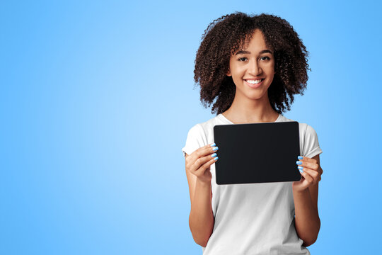 Reviews, offers and online application. Happy young african american female with snow white smile in pink t-shirt with curly hair, shows tablet with blank screen, isolated on purple background