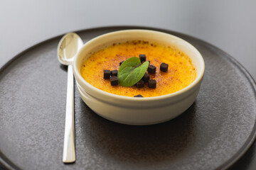 A creamy creme brulee, topped with chocolate and a fresh mint leaf. A delicious dessert on a dark plate with a silver spoon, ready to be savored.