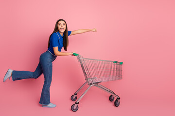Smiling young woman posing casually with a shopping cart on a vibrant pink background, conveying a...
