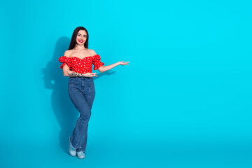 Young woman in red stylish top and blue jeans gesturing against vibrant blue background expressing...