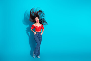 Cheerful young woman tossing hair wearing a red top and jeans against blue background, expressing...