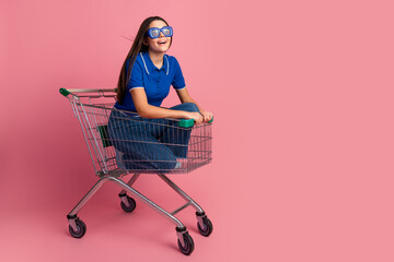 Cheerful young woman sitting in a shopping cart on a colorful pink background showcasing vibrant...