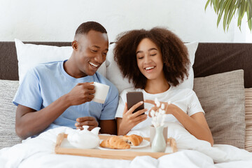 Read news, social networks, great offer and ad at breakfast in bed. Happy millennial african american couple holding cups and drink tea, eat croissants and watch funny video in phone, in bedroom