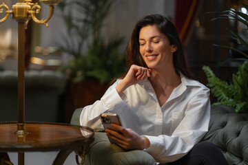 Smiling young businesswoman using smartphone sitting on cushioned chair in hotel lobby. Woman in...