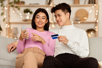 Mobile Banking App. Asian Couple Shopping Online With Smartphone And Credit Card Choosing And Buying Things In Internet Shop On Phone Sitting On Sofa In Cozy Living Room At Home. Selective Focus