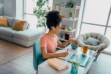 Young woman working from home in a comfortable living space, multitasking with a snack, studies