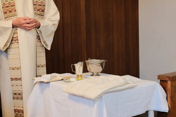 catholic priest in liturgical robe standing beside altar table prepared for eucharist with chalice...