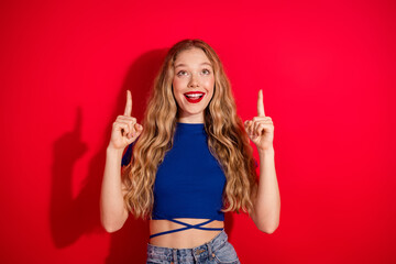 Smiling young woman in a blue top pointing upwards against vibrant red background, expressing...