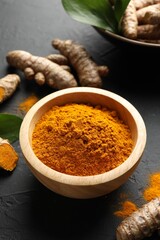 Aromatic turmeric powder in bowl, fresh roots and leaves on black table, closeup