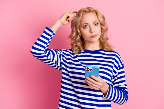 Young Woman in Striped Pullover Scratching Head with Questioning Look Holding Smartphone on Pink Background