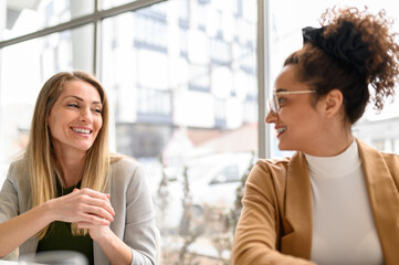 Female colleagues talking and smiling at each other while working together against window in modern...