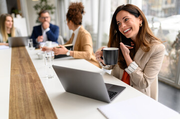 Smiling businesswoman holding coffee cup and talking over smart phone while working with colleagues...