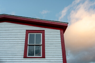 A white narrow wooden clapboard cape cod siding exterior corner wall of a house with bright red trim. A closed glass double-hung window with red wood trim.  Blue sky and clouds in the background.