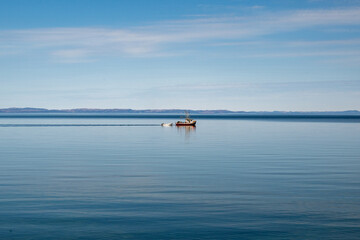 A small fishing boat moves across the calm blue ocean with a colorful blue sky. There are thin clouds over a mountain landmark. The red fishing boat is towing a small white open rowboat or skiff.  © Dolores  Harvey