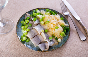 On table is plate with appetizer - pieces of salted herring fillet garnished stewed cabbage salad, decorated spring onion. Gentle refection for alcoholic beverages. Fish supplemented with edible green