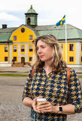 A woman with dark blonde hair stands with a coffee cup, adorned with stylish nails, wedding ring, and modern watch, in front of a historic Swedish yellow building