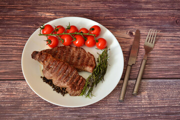 Two grilled beef tenderloin medallions with cherry tomatoes and rosemary on a dark wooden table.
