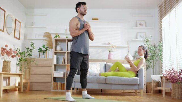 Young asian man doing squat exercise on yoga mat in living room at home while woman using phone for relax on sofa, fitness training and healthy lifestyle in a cozy, strength and strong of muscle.