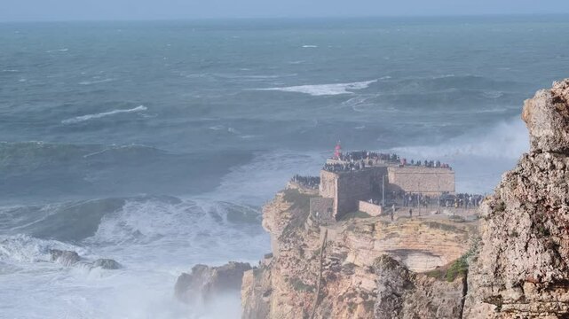 Massive, thundering waves pummeling rugged coastline of nazare, portugal, with crowds gathering at scenic viewpoint to witness powerful oceanic spectacle