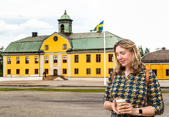 A woman with dark blonde hair smiles down at her coffee cup while standing in front of a charming old yellow building in Sweden, adorned with the Swedish flag
