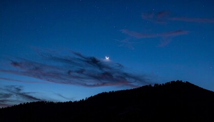 Sliver of moon behind drifting clouds over mountain silhouette at night