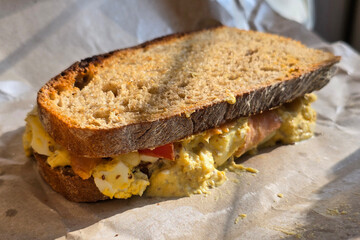 Close-up of a freshly made egg sandwich with tomato slices and creamy filling between toasted whole grain bread, captured in natural morning light.