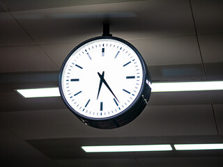 A round, illuminated analog wall clock showing the time, mounted on a ceiling indoors with a modern...