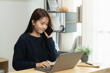 Young Asian woman in casual sweater talking on smartphone while working on laptop at home office, creating productive environment with greenery and natural light