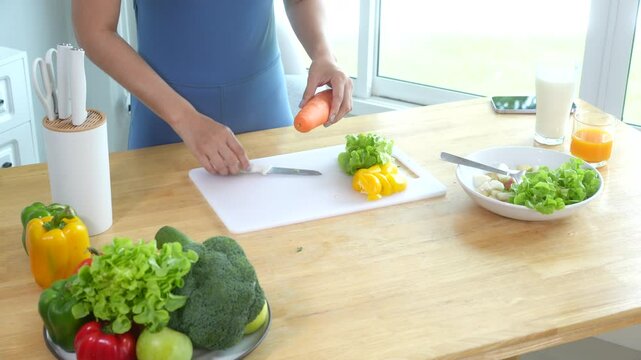 Beautiful Asian woman in fitness sportswear happily smiles while slicing vegetables and fruits on cutting board.She focuses on health nutrition at home,offering online guidance through her computer