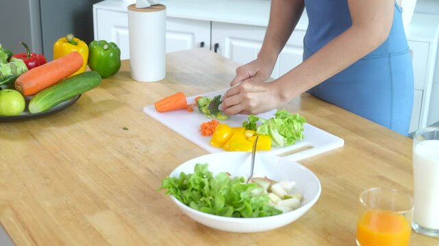 Beautiful Asian woman in fitness sportswear happily smiles while slicing vegetables and fruits on cutting board.She focuses on health nutrition at home,offering online guidance through her computer