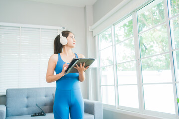 A focused woman in blue activewear intently uses a tablet, possibly for a workout routine, in a bright, modern home setting with a soft grey couch and natural light filtering through nearby blinds