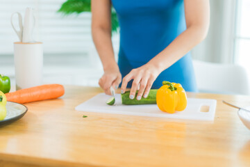 Beautiful Asian woman in fitness sportswear happily smiles while slicing vegetables and fruits on cutting board.She focuses on health nutrition at home,offering online guidance through her computer