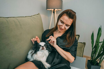 woman in black robe with under eye patches gently petting sleepy white and black japanese chin dog on green sofa in cozy home lamp and houseplant, wellness, pets care, lifestyle brands
