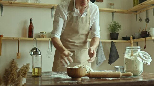 A baker in an apron kneads the dough with his hands and sprinkles it with flour to bake delicious homemade bread.