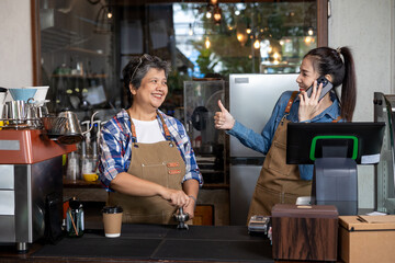 Asian senior woman and young Asian woman working together inside cozy coffee shop wearing aprons smiling with happiness while one talks on smartphone and gives thumbs up showing teamwork 