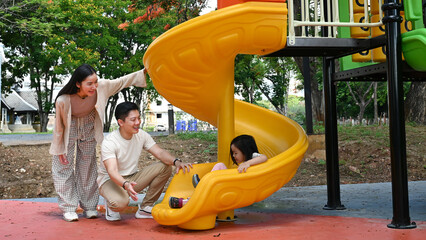 A family is standing in front of a slide. The child is sitting on the slide while the parents look on. Scene is happy and playful.